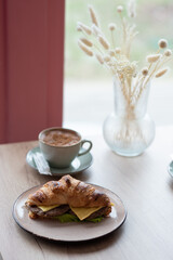 Close-up of a mug with coffee and a croissant with cheese and ham standing on a table in a cafe, near the window.