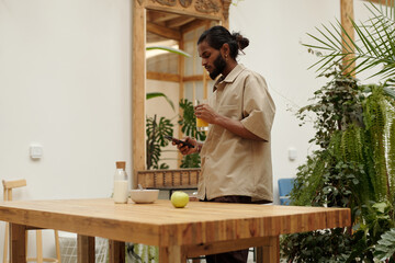 Architect with long hair tied back drinking juice while checking his phone at table with green plant surroundings