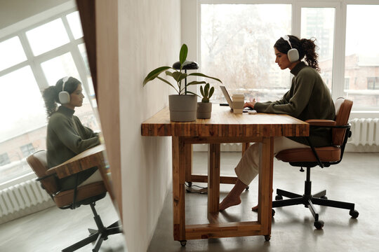 Architect seated at wooden desk using laptop, wearing headphones, and viewing soft window light