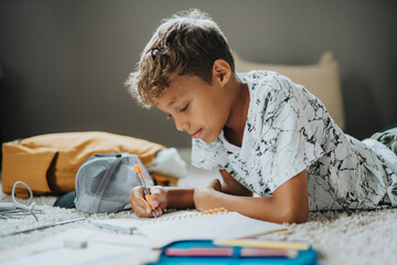 Focused boy writing in diary while lying on front at home