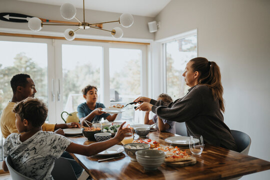 Mother giving pizza slice to son sitting near dining table for lunch at home