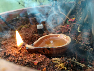 A traditional clay diya with a small flame sits on a bed of soil, surrounded by wisps of incense smoke. The warm, golden light contrasts beautifully with the earthy tones of the surrounding space.