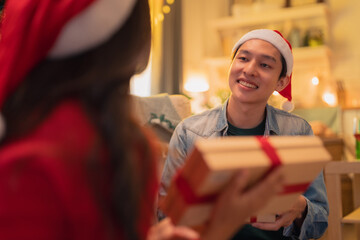 A joyful man in Santa hat smiles as he receives beautifully wrapped gift from woman in festive attire, capturing warmth of holiday spirit