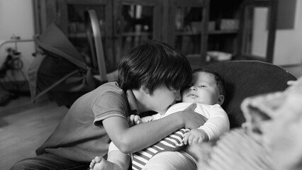 Black and white shot of older sibling kissing baby’s cheek while baby sits in a bouncer, a tender and loving moment between siblings in a peaceful home environment