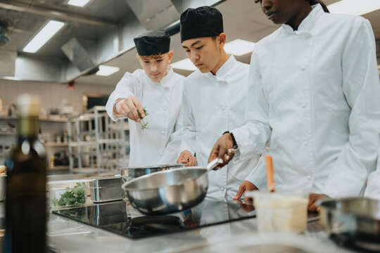Male teenage trainees preparing food by female colleague holding saucepan in commercial kitchen - Powered by Adobe
