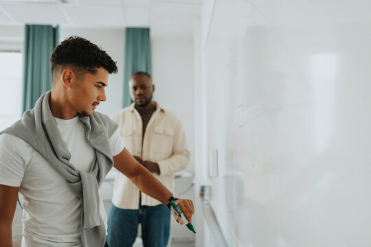 Teenage student with felt tip pen writing on whiteboard with teacher in background