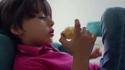 Close-up of young boy holding and eating an apple while lying back on a couch, child in a relaxed and quiet moment of eating and contemplation