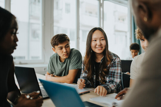 Smiling teenage girl discussing with male teacher while sitting with friends in classroom