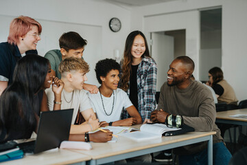 Happy male and female students with non-binary person discussing with teacher sitting at desk in classroom