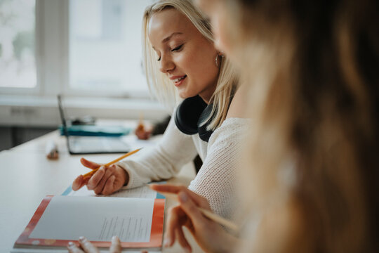Blond teenage girl reading book while discussing with female teacher in classroom