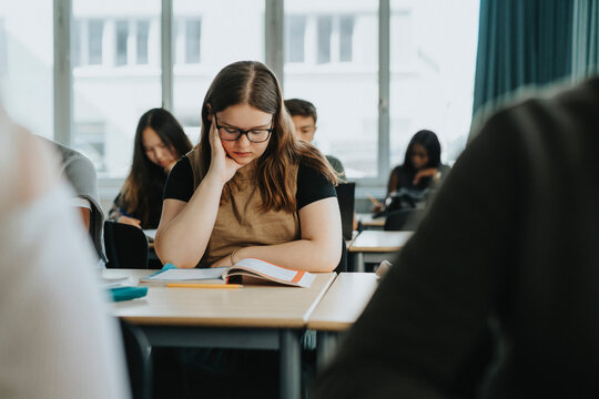 Teenage girl with head in hand reading book while sitting at desk in classroom