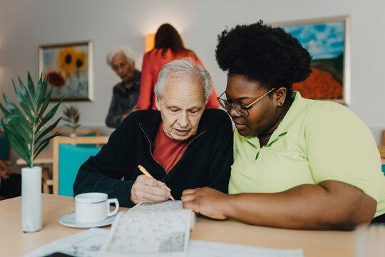 Nurse sitting next to senior man solving Sudoku puzzle at retirement home
