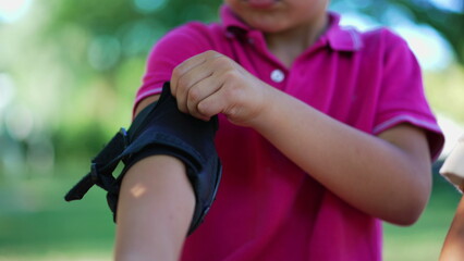Young boy fastening elbow pad, focused on adjusting protective gear before playing, showing concentration and attention to safety for outdoor activities in a park setting