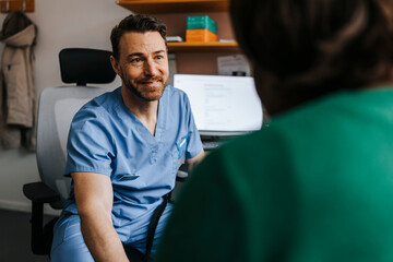 Smiling medical professional in scrubs consoling patient while sitting on chair in examination room
