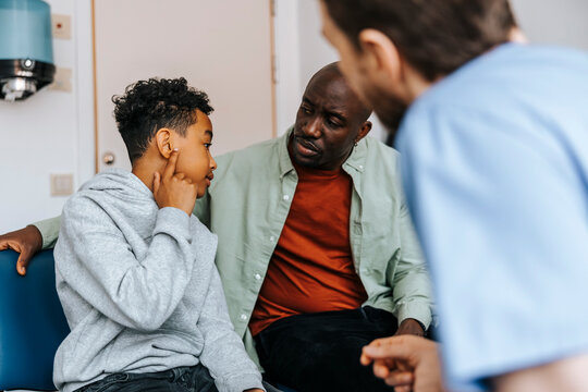 Boy showing ear problem to doctor while sitting next to father in medical examination room