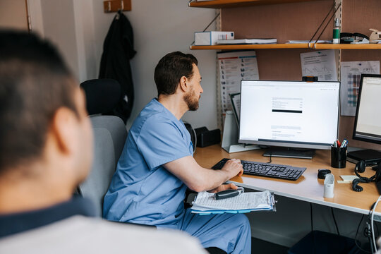 Male doctor checking medical records of patient on computer while sitting near table at medical examination room