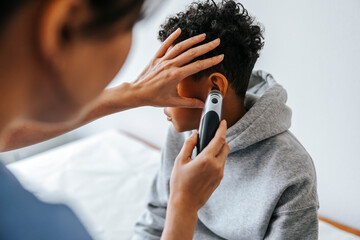 Female pediatrician checking boy's ear with thermometer in medical examination room