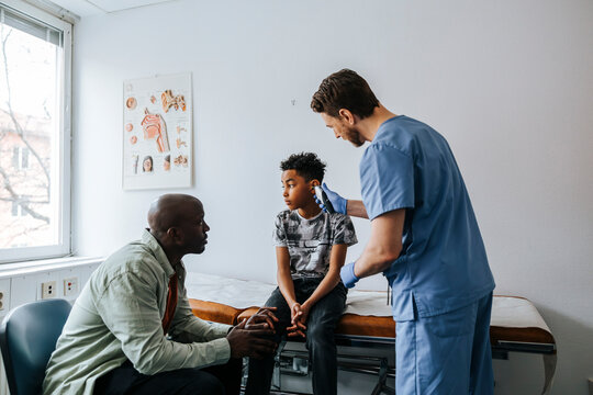 Male pediatrician examining boy's ear with thermometer while father sitting on chair in examination room