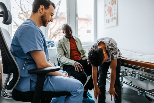 Side view of male doctor asking boy to bend over for medical exam while sitting in examination room