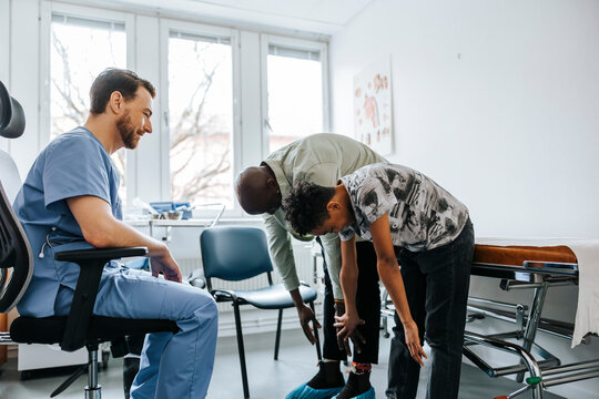 Side view of male doctor asking father and son to bend over while sitting in medical examination room
