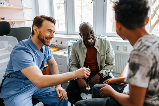 Smiling male healthcare worker holding reflex hammer while sitting with father and son during visit in examination room