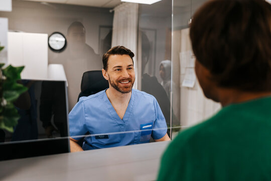 Happy male receptionist seen through glass assisting patient standing at medical clinic
