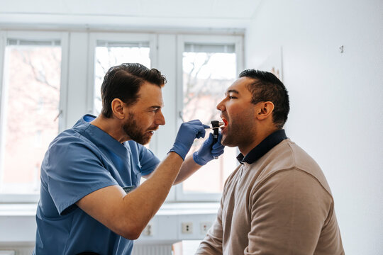 Male medical expert examining throat of man using tongue depressor in examination room at hospital