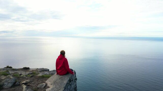 Girl in red jacket sitting on top of mountain with beautiful view of norwegian sea, Ryten, Lofoten islands