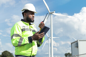 African engineer with safety vest and helmet holds walkie talkie and tablet at wind turbine farm....