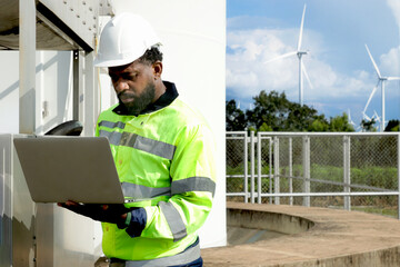 African engineer with safety vest and helmet working on laptop computer at wind turbine farm. Worker works at windmill park with cloudy sky. Engineering ecology alternative renewable energy technology