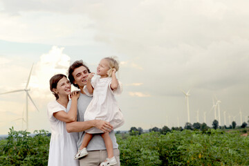 Naklejka premium Happy family at wind turbine farm. Mother and father who carry daughter spending time together at sunset sky windmill park. Parents and girl kid take a walk, ecology alternative renewable energy.