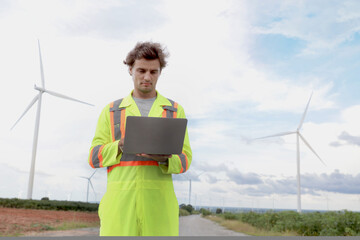 Young engineer with safety vest working on laptop computer at wind turbine farm. Worker works at windmill park with cloudy sky. Engineering and ecology alternative renewable energy technology concept.