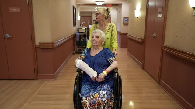 Family daughter visiting, pushing mother with broken arm in wheelchair in rehab facility.