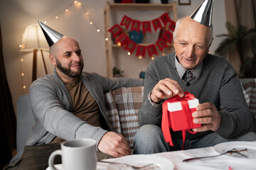 Happy smiling adult son giving surprise gift to elderly father while sitting on sofa. concept of birthday and bonding