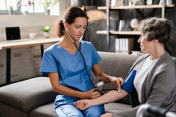 Middle-aged nurse caregiver doctor medical worker checking pulse and blood pressure of old elderly...