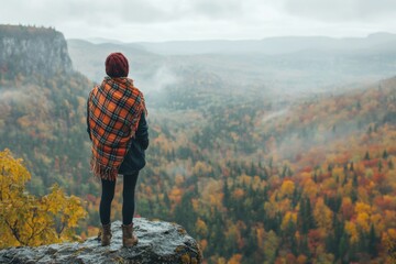 Naklejka premium solo travel adventure - person standing on a cliff edge, overlooking a valley filled with autumn colors, wrapped in a plaid scarf, minimal background with copy space
