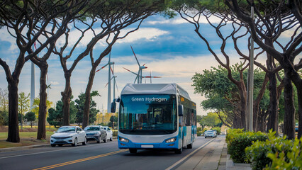 A hydrogen-powered public bus with a Green Hydrogen label, cruising down a tree-lined street with wind turbines visible in the background.