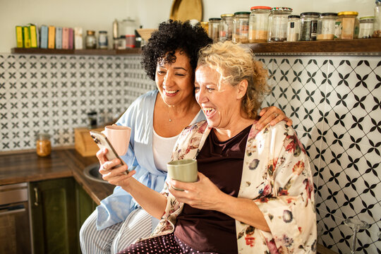 Happy mature lesbian couple laughing together while looking at phone and drinking coffee in kitchen - Powered by Adobe