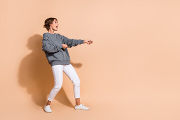 Full length photo of impressed girl wear pullover white trousers pull object on rope from empty space isolated on beige color background