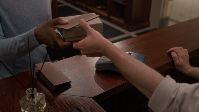 Close up view of unrecognizable Black customer paying by credit card and receiving small wrapped package from cashier at wooden counter with bottle of fragrance sticks in boutique