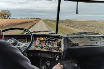 Inside View of an Old Semi Truck Dashboard: Dusty and Worn with Use, Featuring the Driver’s Hand on the Steering Wheel as He Shifts Gears, Driving on a Rural Dirt Road with Corn Crops and a Blue Sky