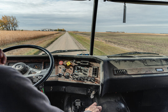 Inside View of an Old Semi Truck Dashboard: Dusty and Worn with Use, Featuring the Driver&rsquo;s Hand on the Steering Wheel as He Shifts Gears, Driving on a Rural Dirt Road with Corn Crops and a Blue Sky