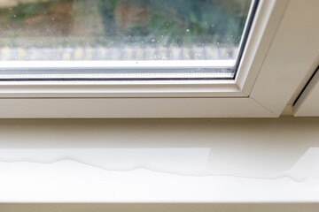 Top view of a white window frame with a window sill, which spreads a puddle. There is a gap in the double-glazed window, the window frame is leaking