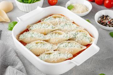 Raw conchiglioni pasta stuffed ricotta cheese and spinach with tomato sauce and parmesan cheese on top in a white baking dish on a gray concrete background.