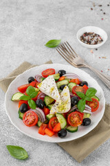 Greek salad of fresh juicy vegetables, feta cheese, herbs and olives on a white plate on a light concrete background. Healthy food. Copy space, top view.