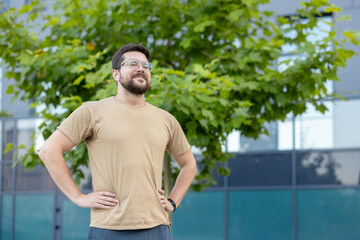 Smiling and relaxed young man resting after jogging and sports training, standing outdoors, holding...