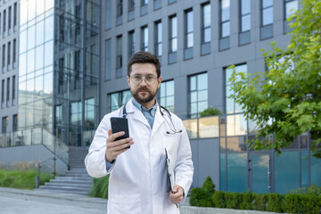 Portrait of a serious young male doctor in a white coat and glasses standing with a folder in his hand near the clinic, holding a mobile phone and looking confidently at the camera