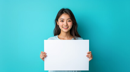 Young smiling asian woman holding a large blank white paper in both hands against a soft pastel blue background.
