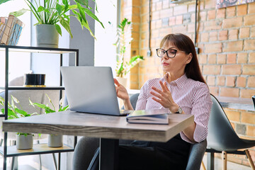 Talking middle-aged woman having video call using laptop in coworking cafe