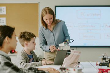 Obraz premium Curious school boy at desk looking at robot while professional teacher showing it